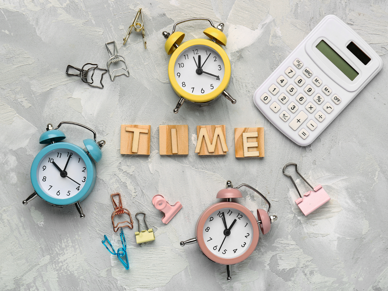 Wooden letter blocks spelling the word “TIME” surrounded by colorful clocks, paper clips, and a calculator.