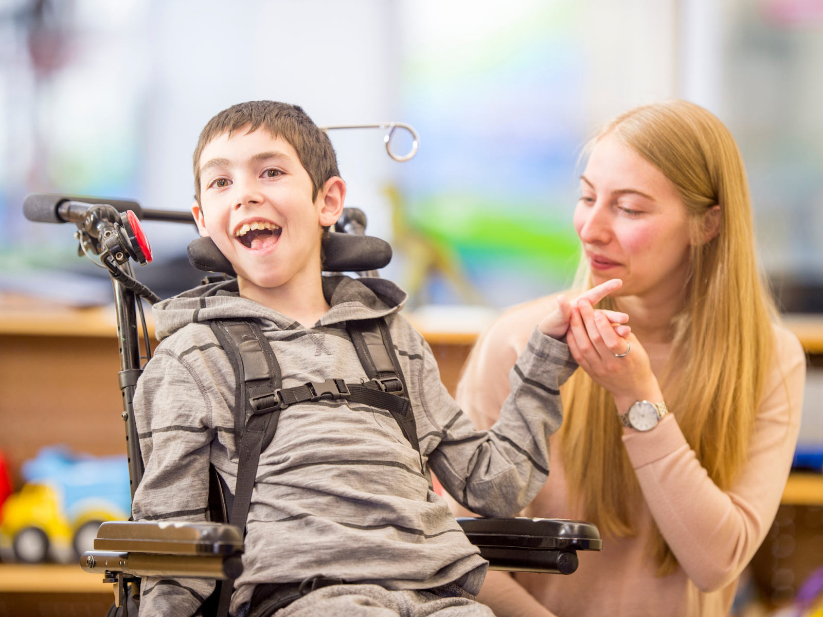 A disability support aid kneeling next to a young smiling boy in a wheelchair.