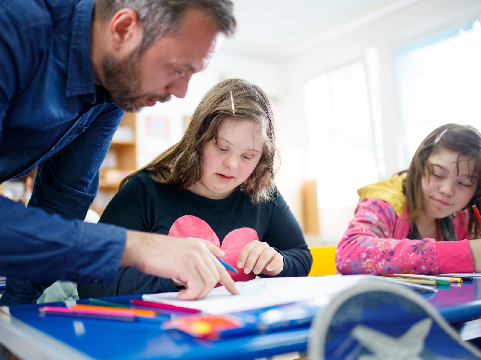 An adult leaning over a desk and gesturing to where a child with a disability is drawing.