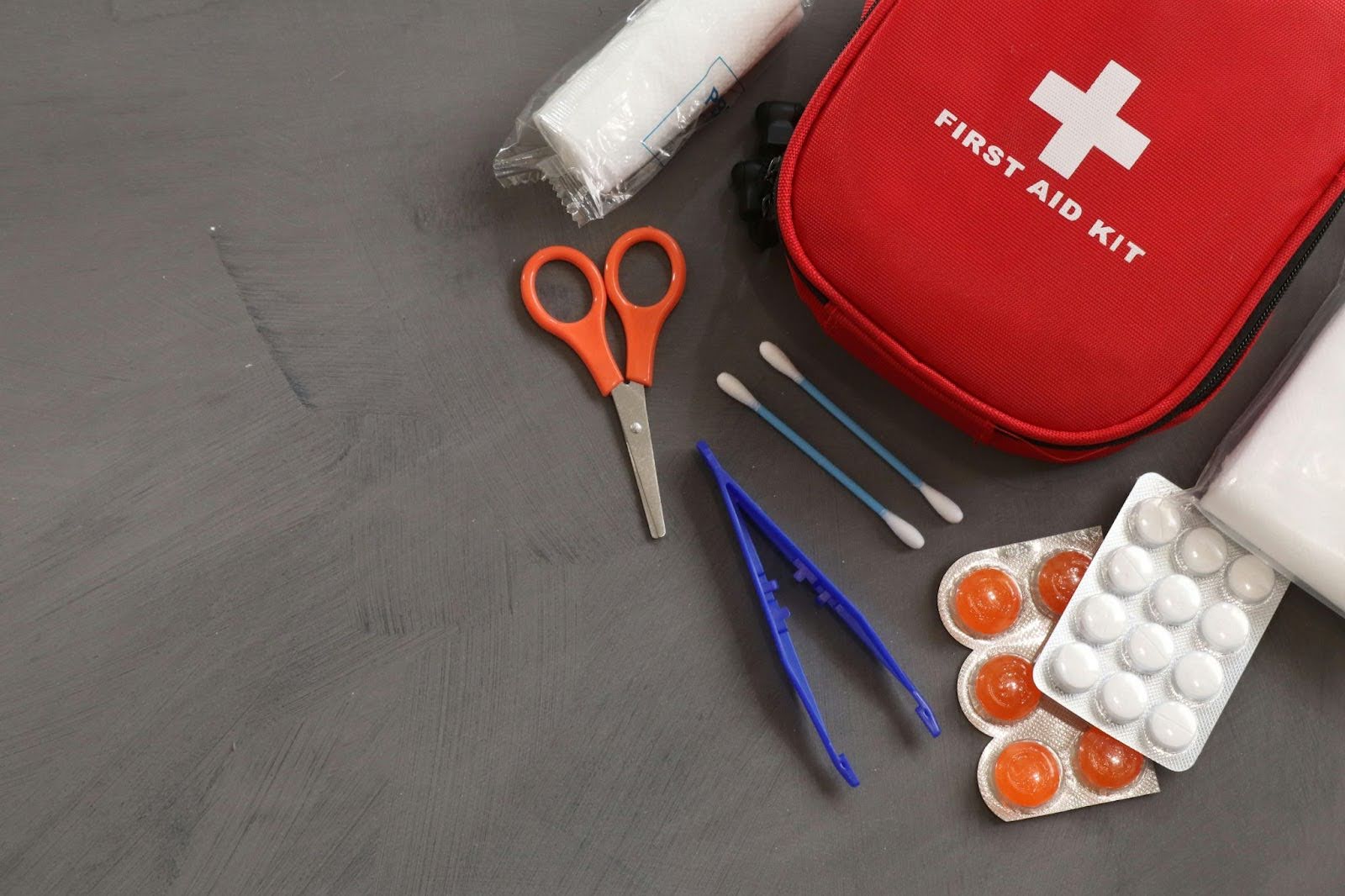 Some items that you might find in an emergency preparedness kit – including a first aid kit, some scissors, medical gauze, tweezers, cotton swabs, and lozenges – sit on top of a gray background