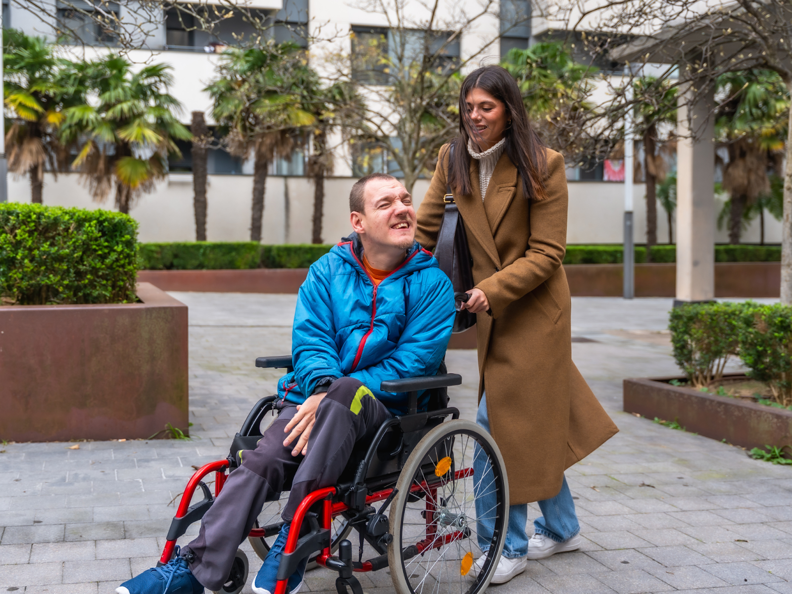 A primary caregiver smiling while pushing a man in a wheelchair through a courtyard.
