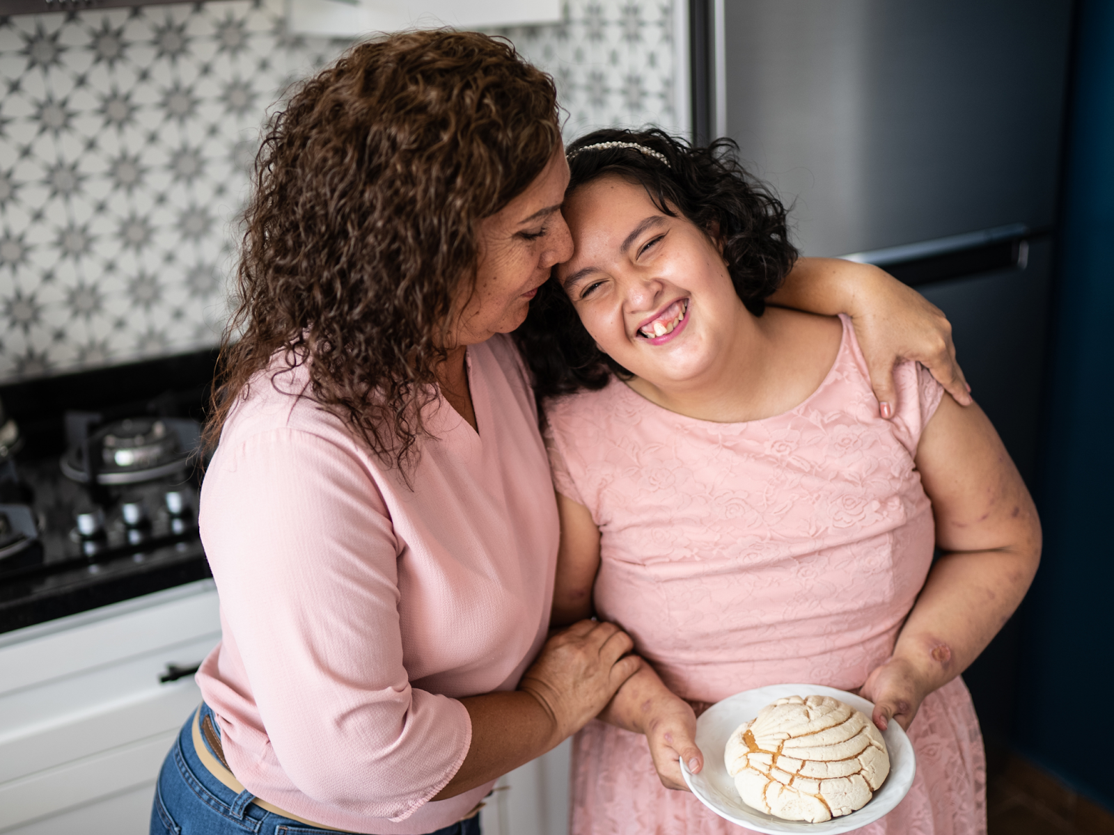 A mother with her arms around her smiling daughter with an intellectual disability, who is holding a plate with a pastry.