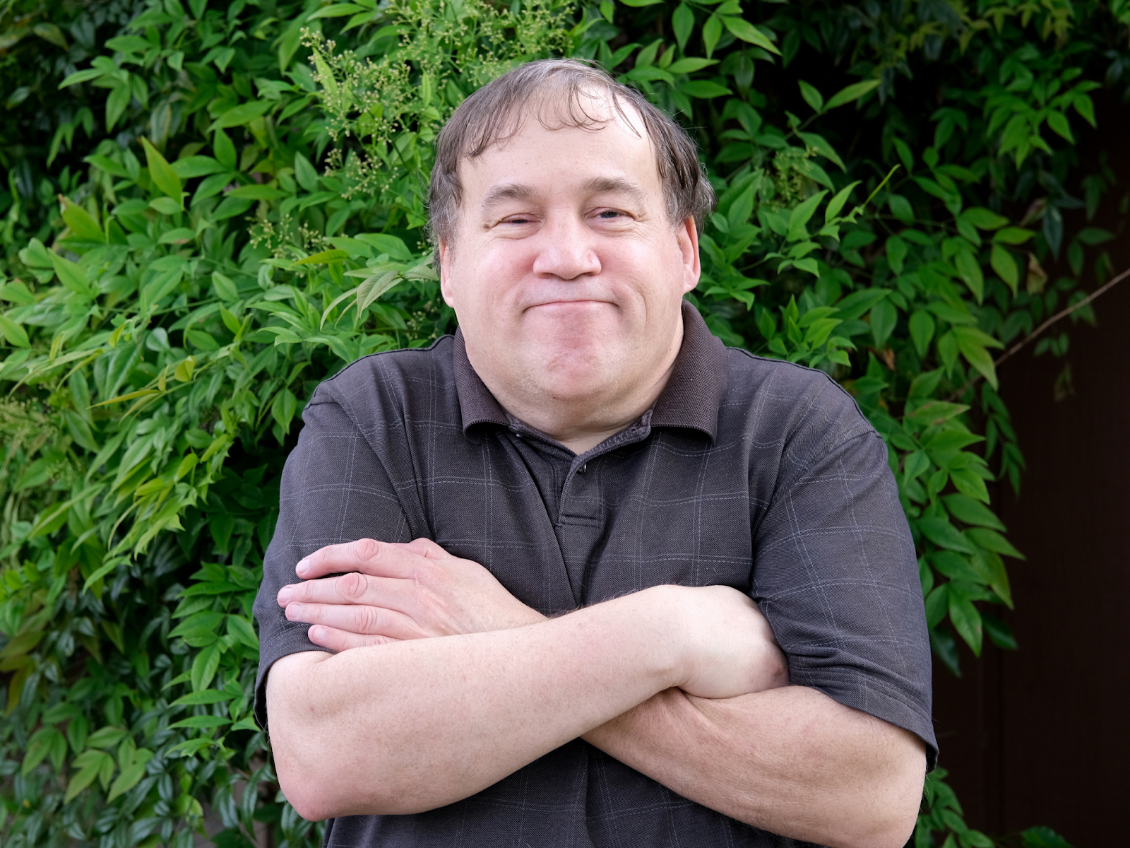An older autistic adult standing in front of greenery and smiling with his arms crossed.
