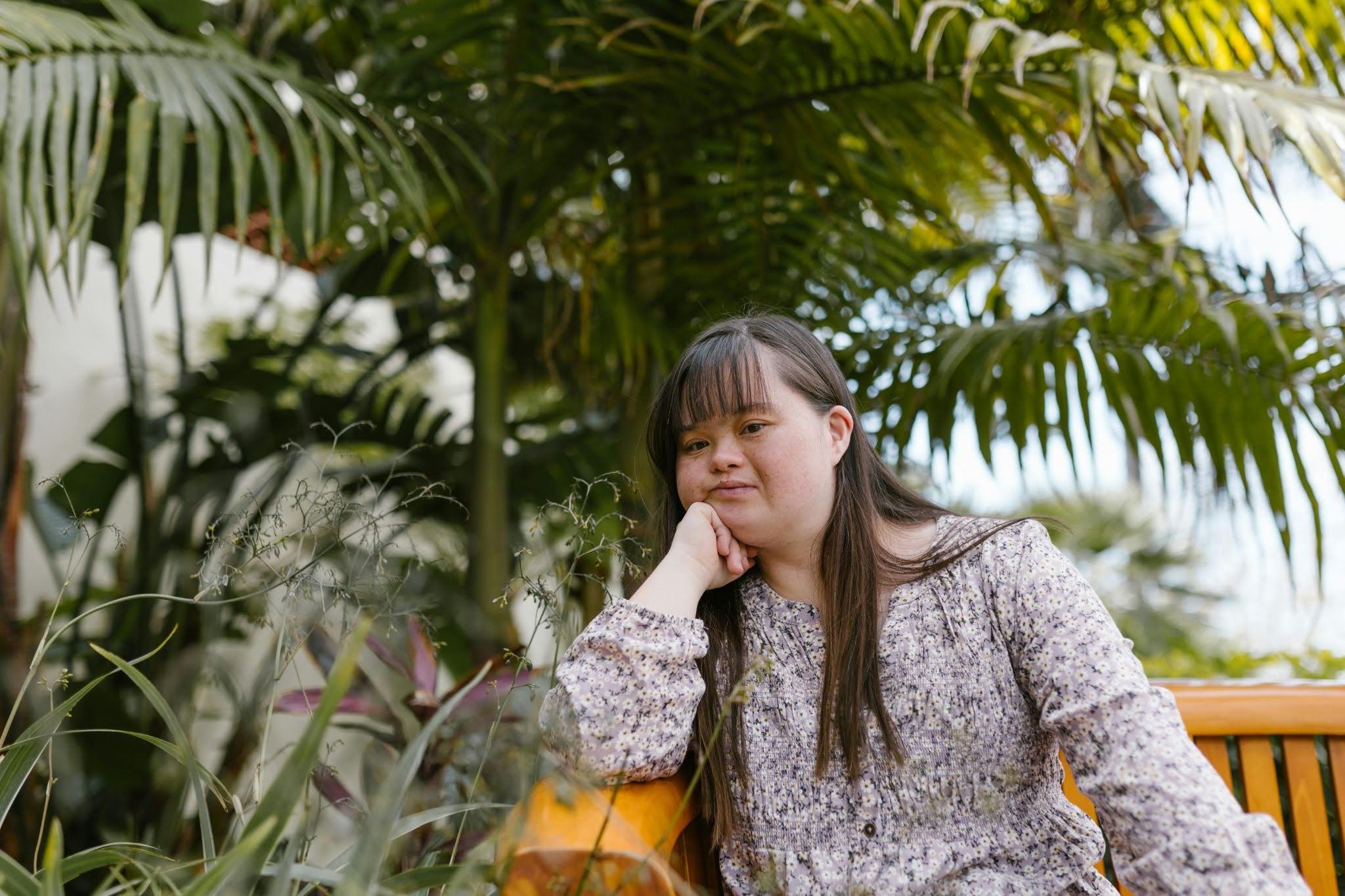 A young woman with Down syndrome sits on a bench next to some trees while resting her head on her hand