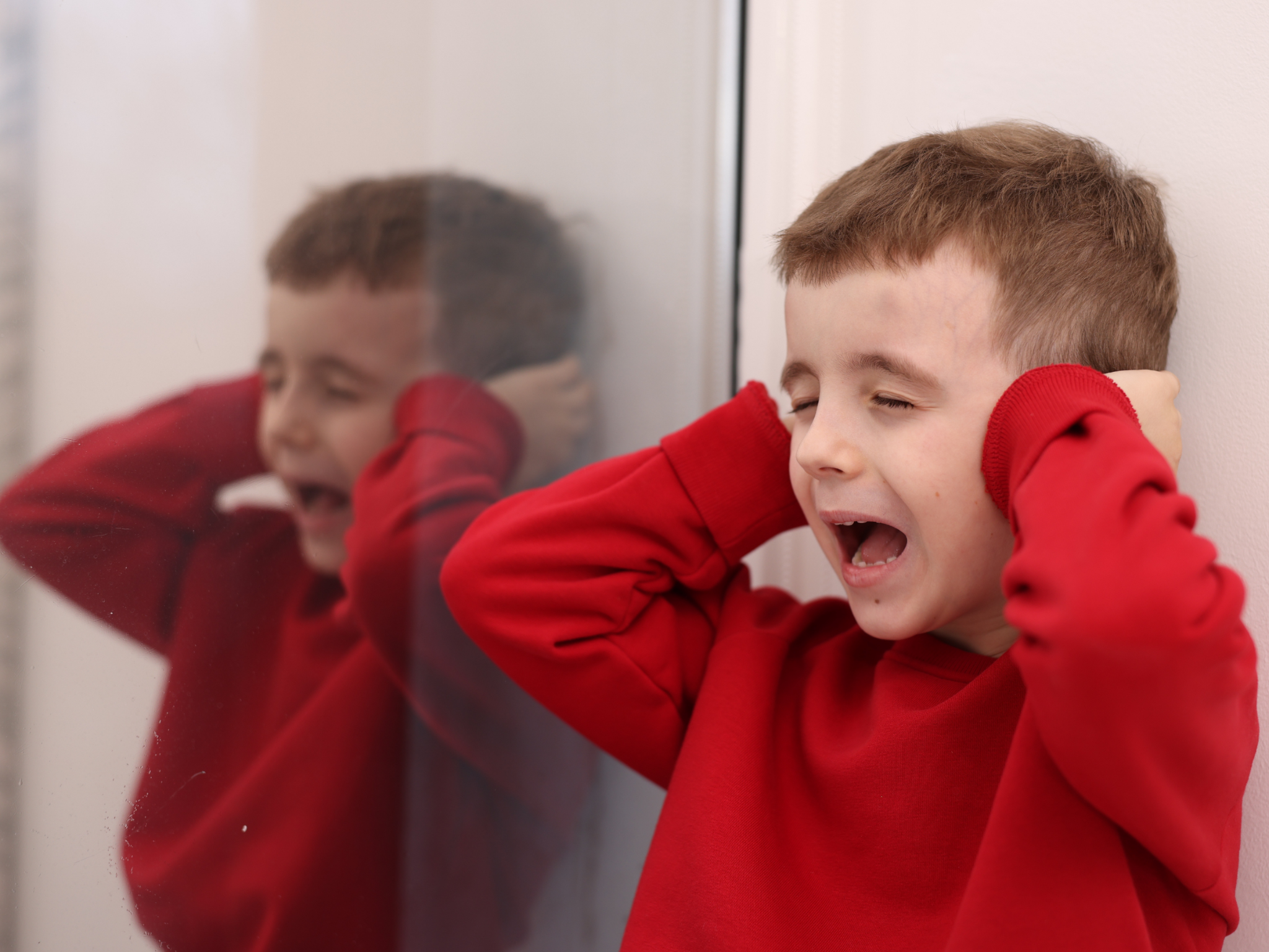 A boy in a red shirt closing his eyes and holding his hands over his ears.