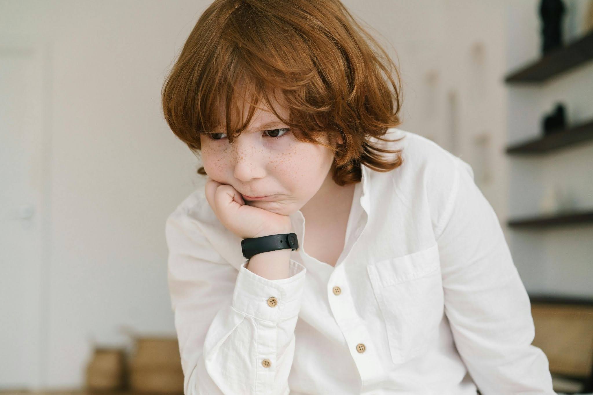A child in a white shirt pouting with their head resting on their hand.