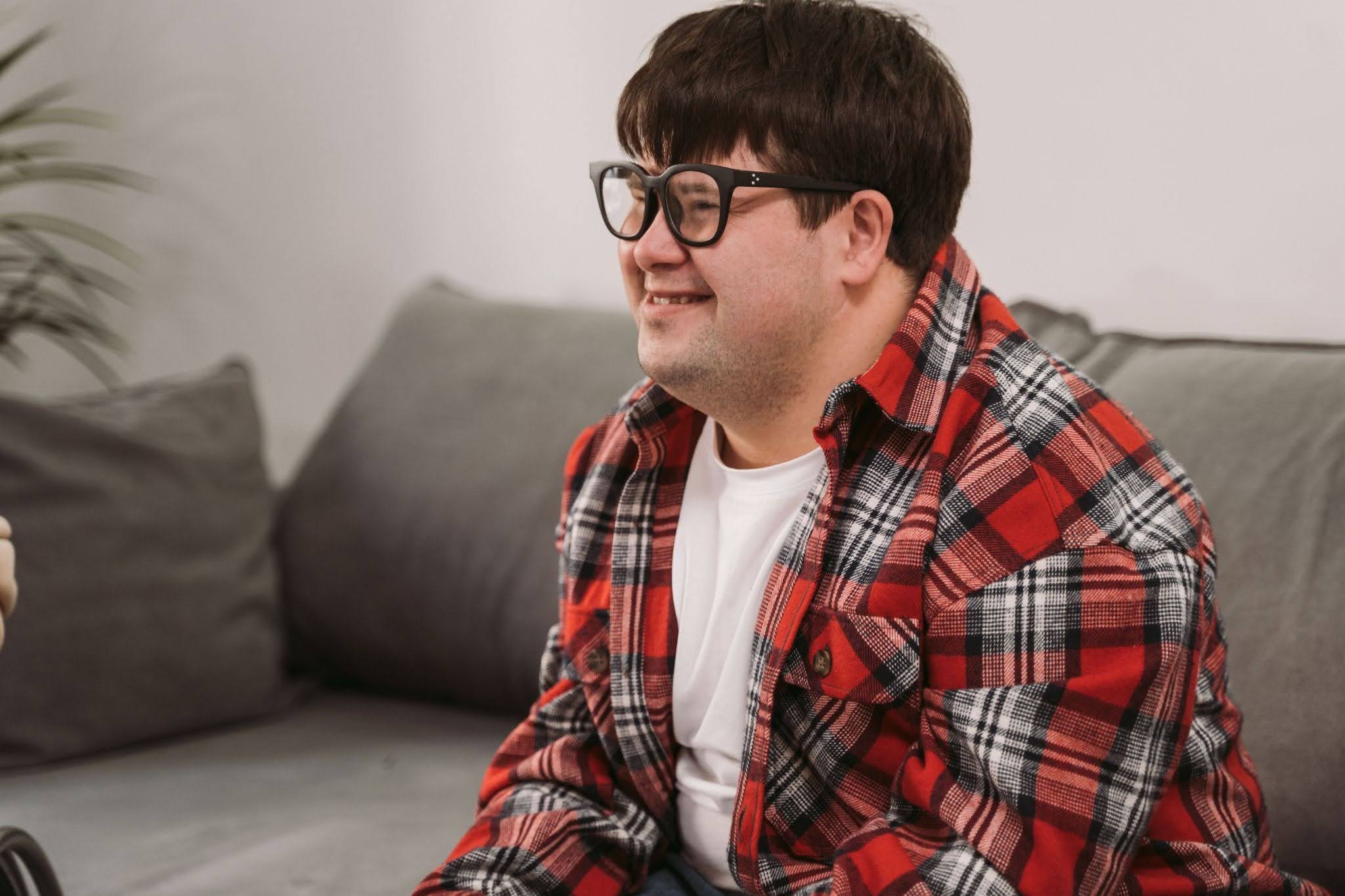 A young man with Down Syndrome wearing glasses sits on a couch and smiles