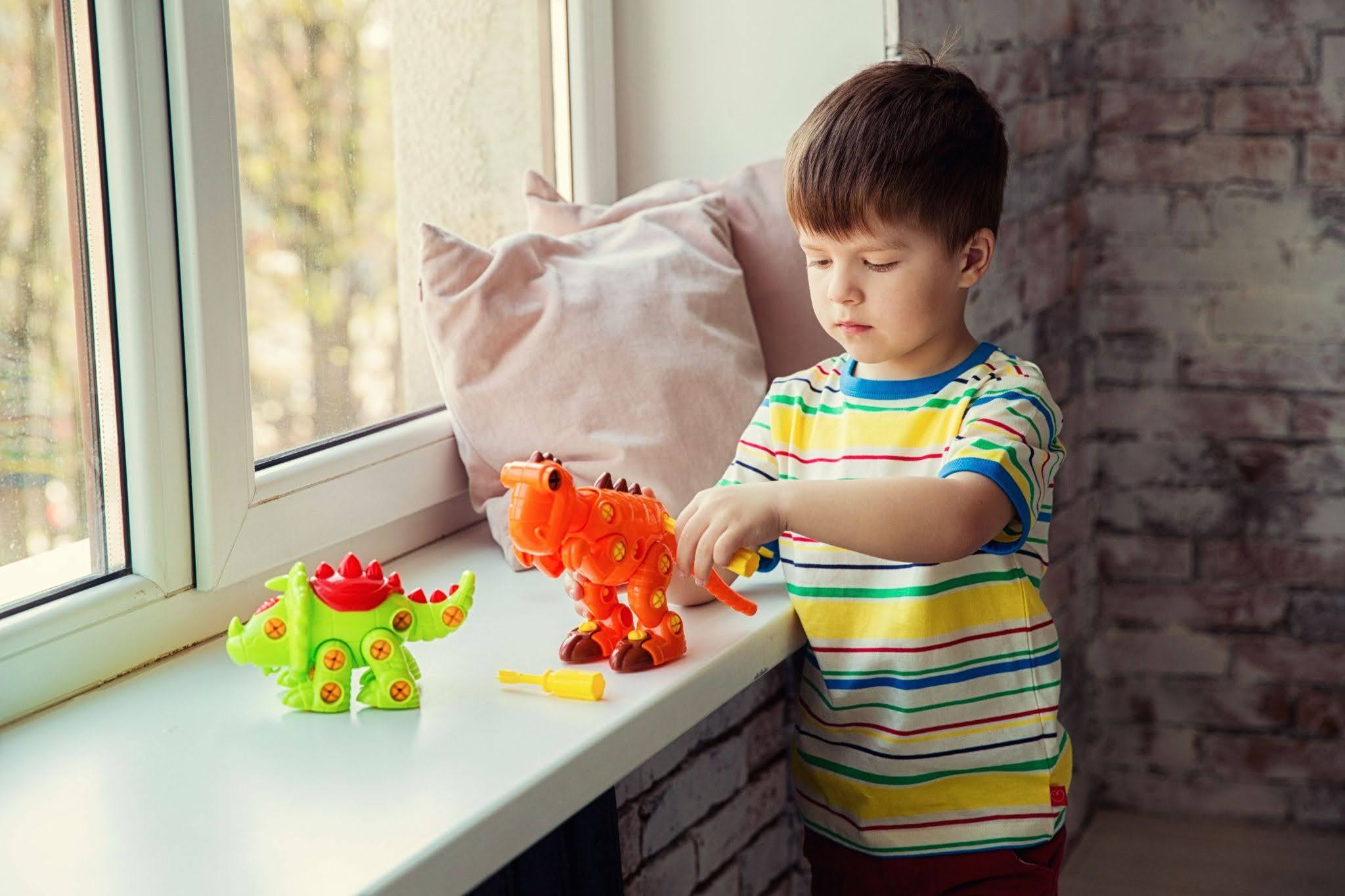 A young child plays with brightly colored green and orange dinosaur toys on a windowsill