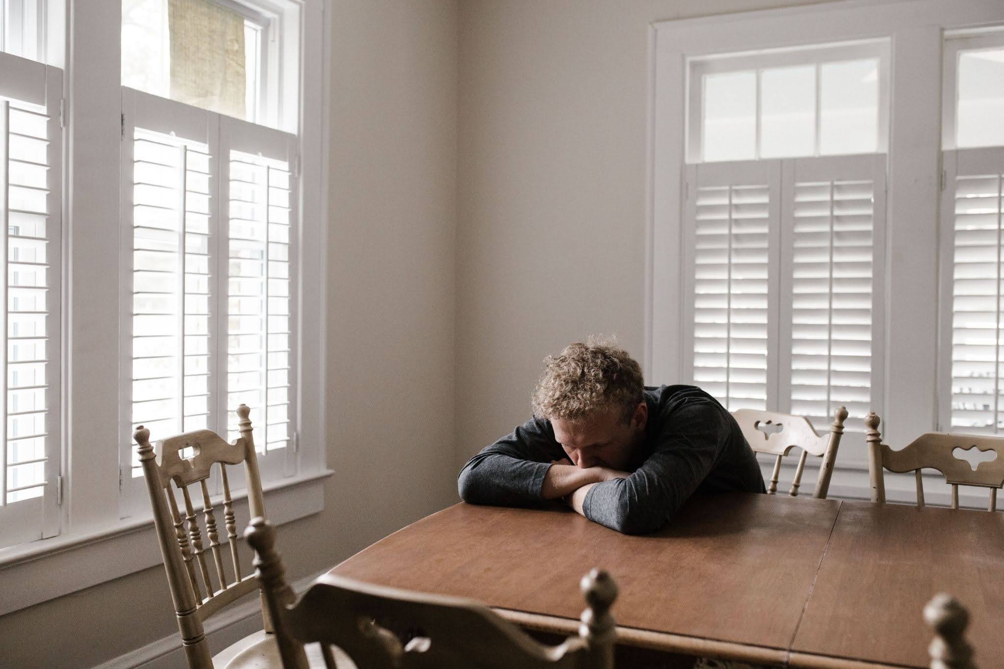  A man experiencing depression sits alone at a kitchen table, his head resting on his folded arms