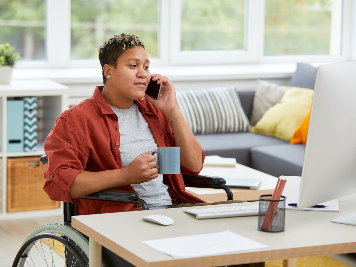 Person at home in a wheelchair talking on a cell phone, facing a desk with a computer