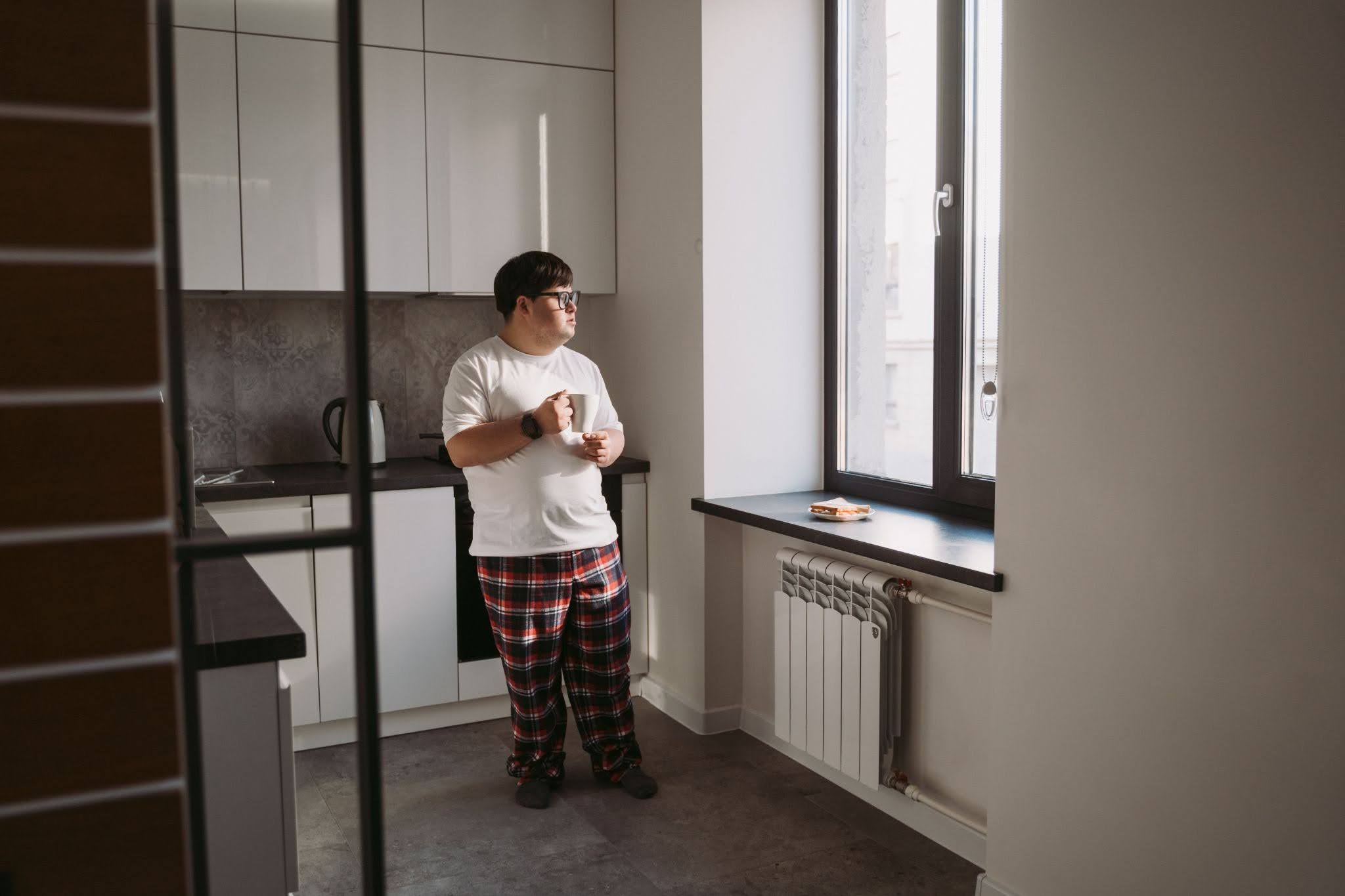 A man stands in his apartment, holding a mug and staring out the window