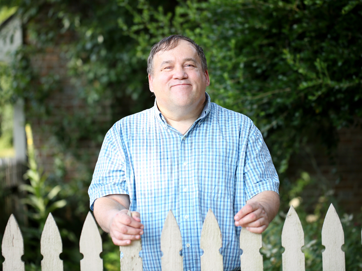 A man stands behind a white picket fence and smiles