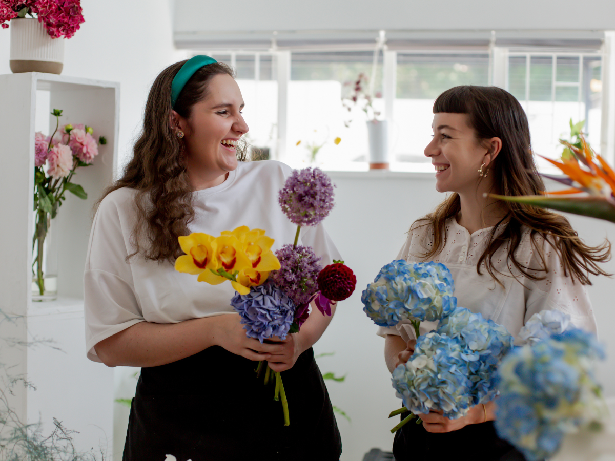 Two women holding flowers, looking at each other and smiling.