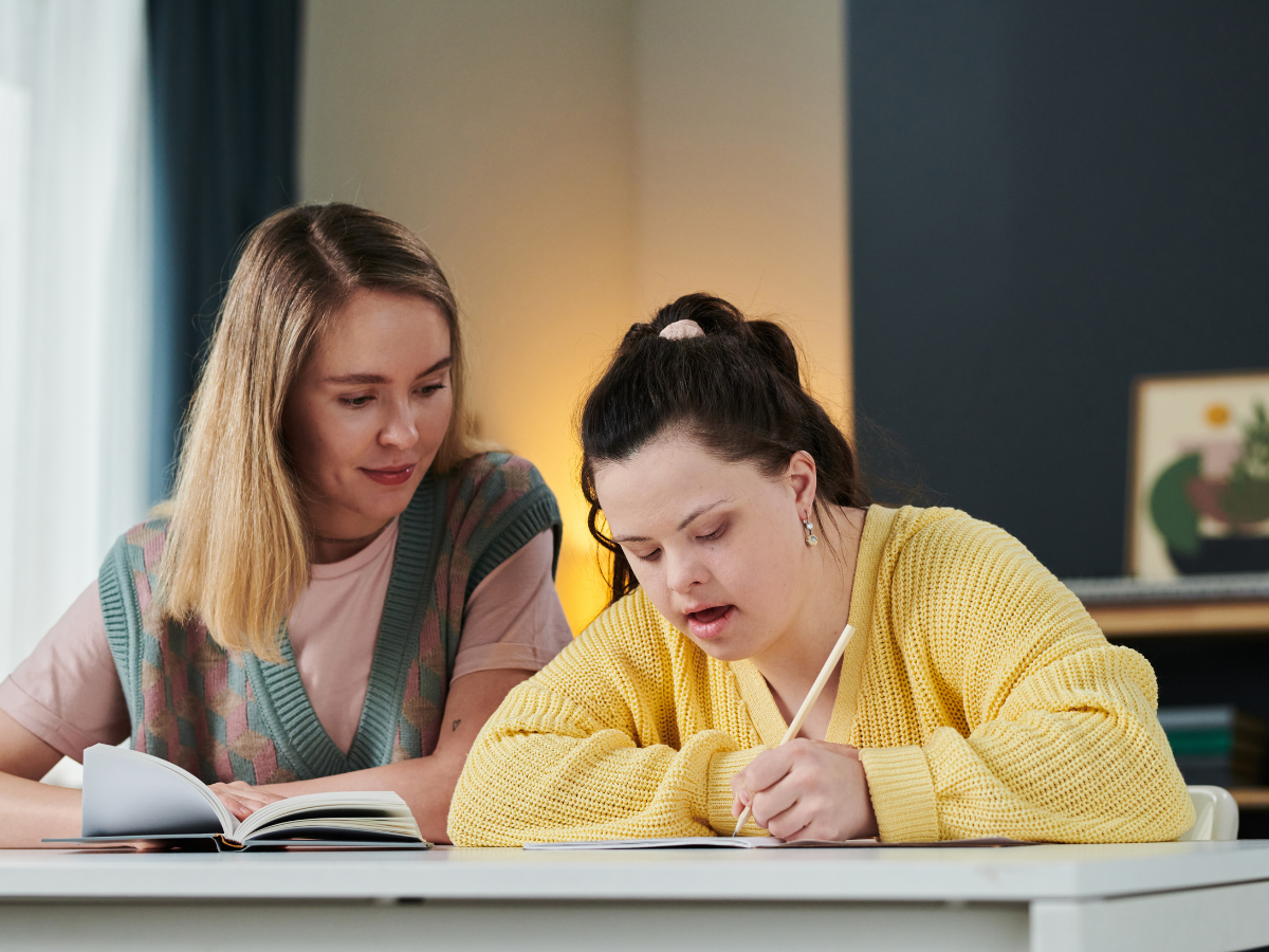 A teacher sits next to a student with Down syndrome writing