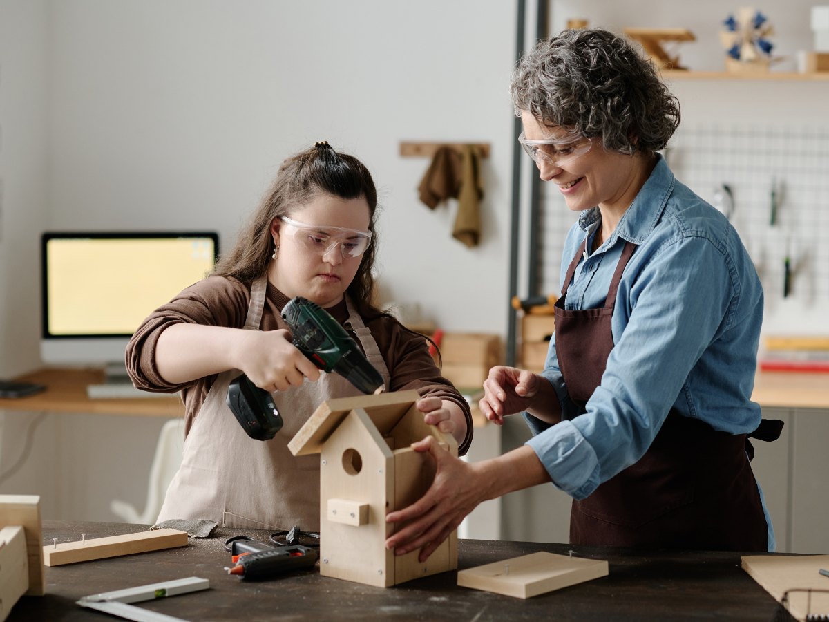 A teacher helping a young adult with a disability build a birdhouse