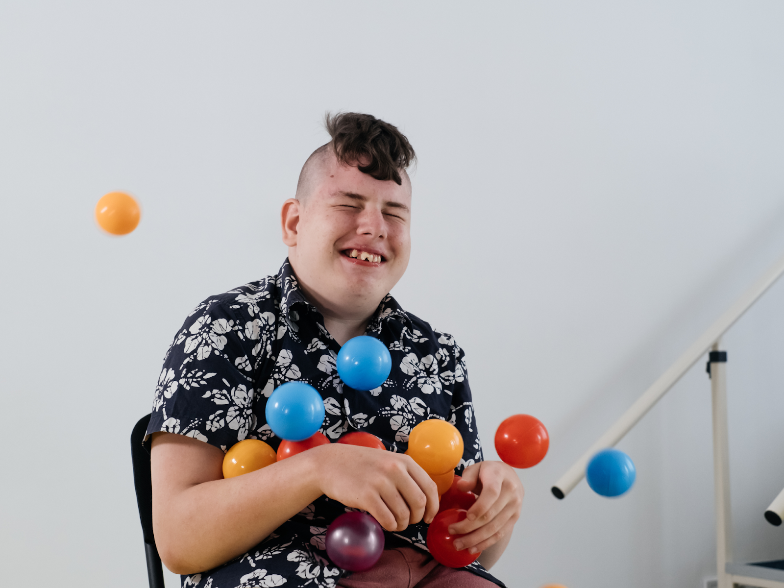 A autistic young adult sits on a chair and smiles while playing with some brightly-colored balls