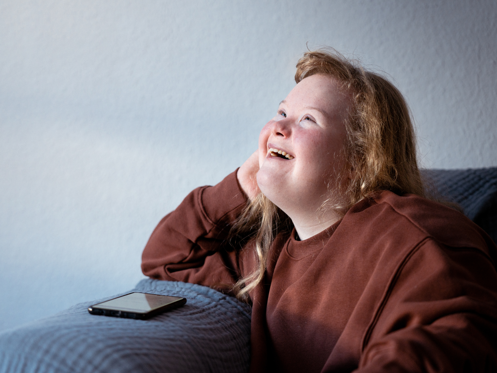 A young woman with Ehlers-Danlos Syndrome sits on a couch and smiles while looking upward