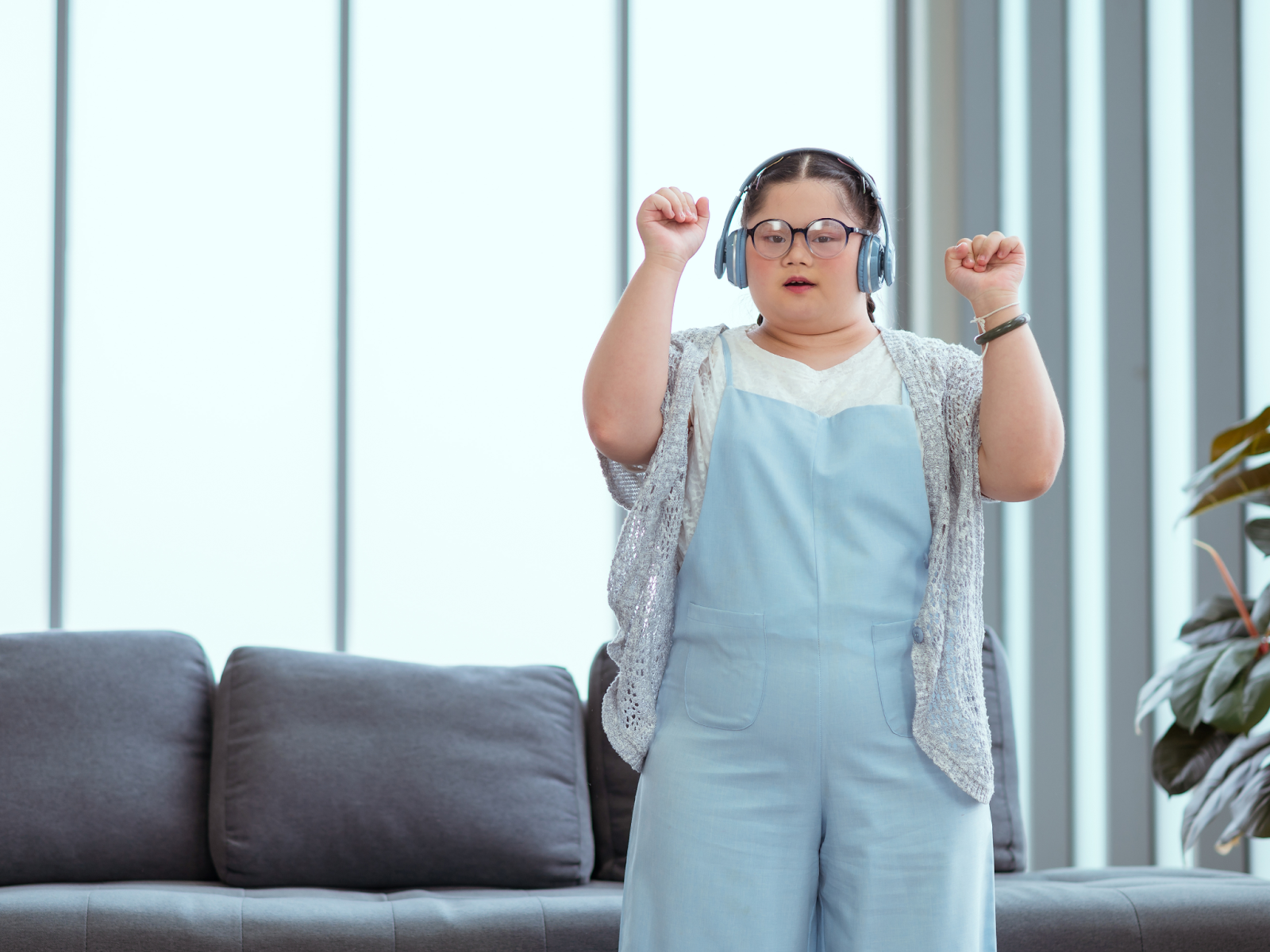 A neurodivergent person wearing glasses and headphones raises both hands while dancing in a well-lit living room. 