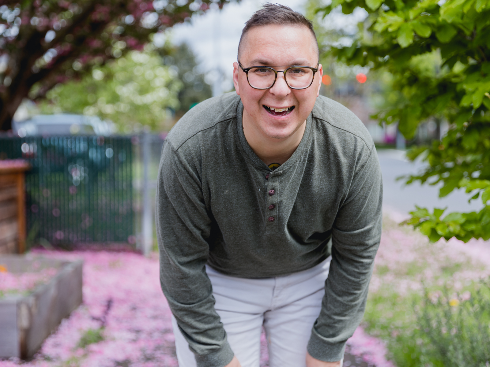An autistic person with glasses leans forward and smiles, surrounded by trees and pink fallen leaves.