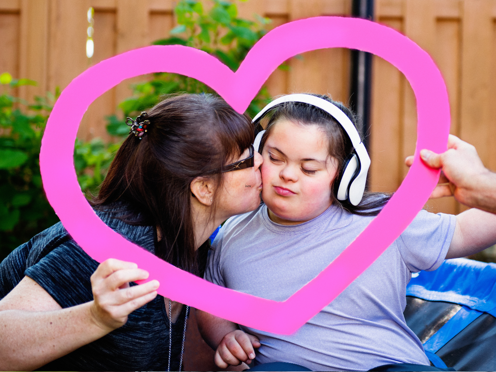 A woman kisses a girl wearing headphones on the cheek with their faces framed in a pink heart cutout