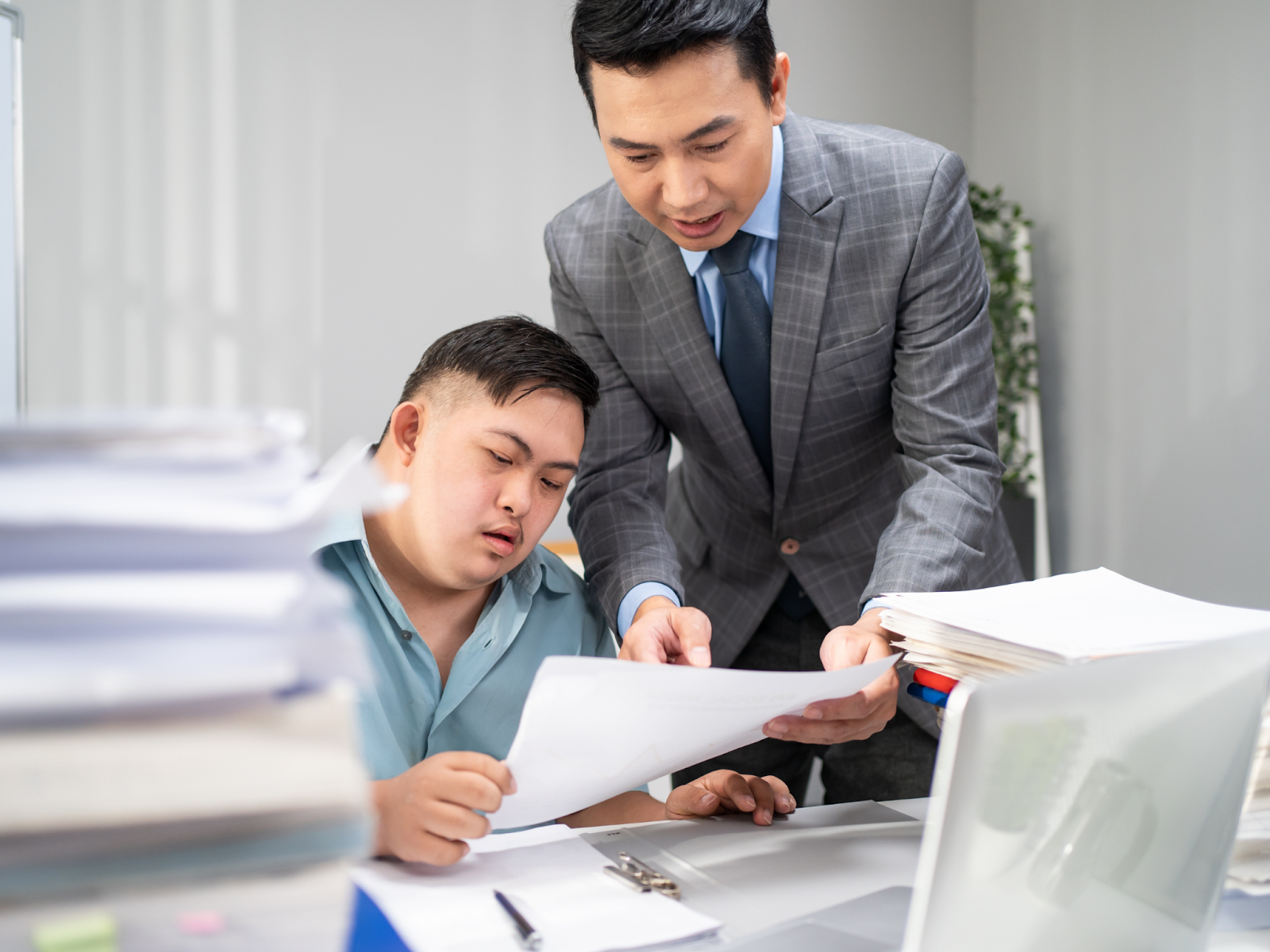 A man in a suit standing over a desk and pointing to a piece of paper with an adult with a disability.
