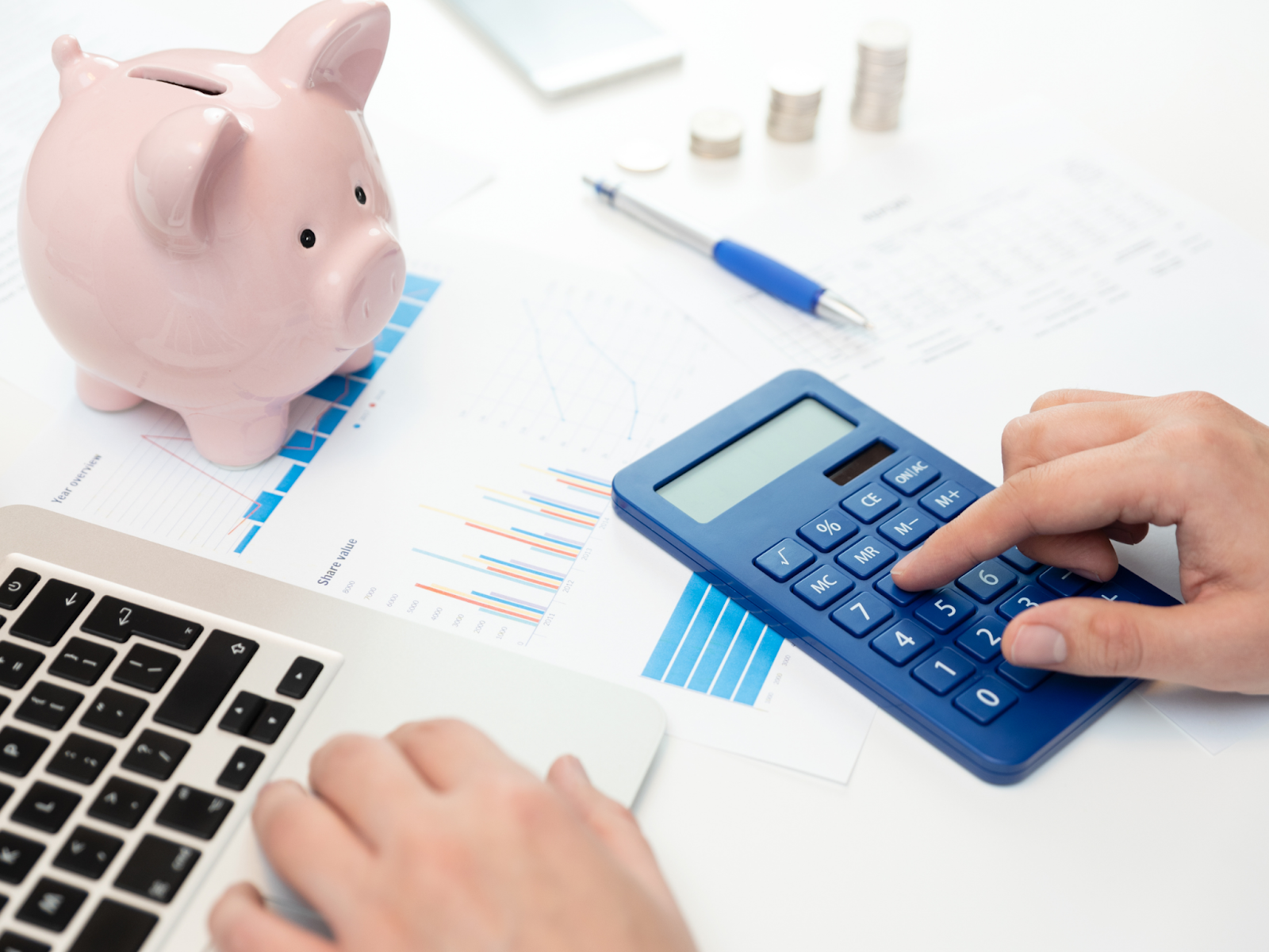 Two hands over a desk crowded with financial papers, stacked coins, a piggy bank, and a calculator.
