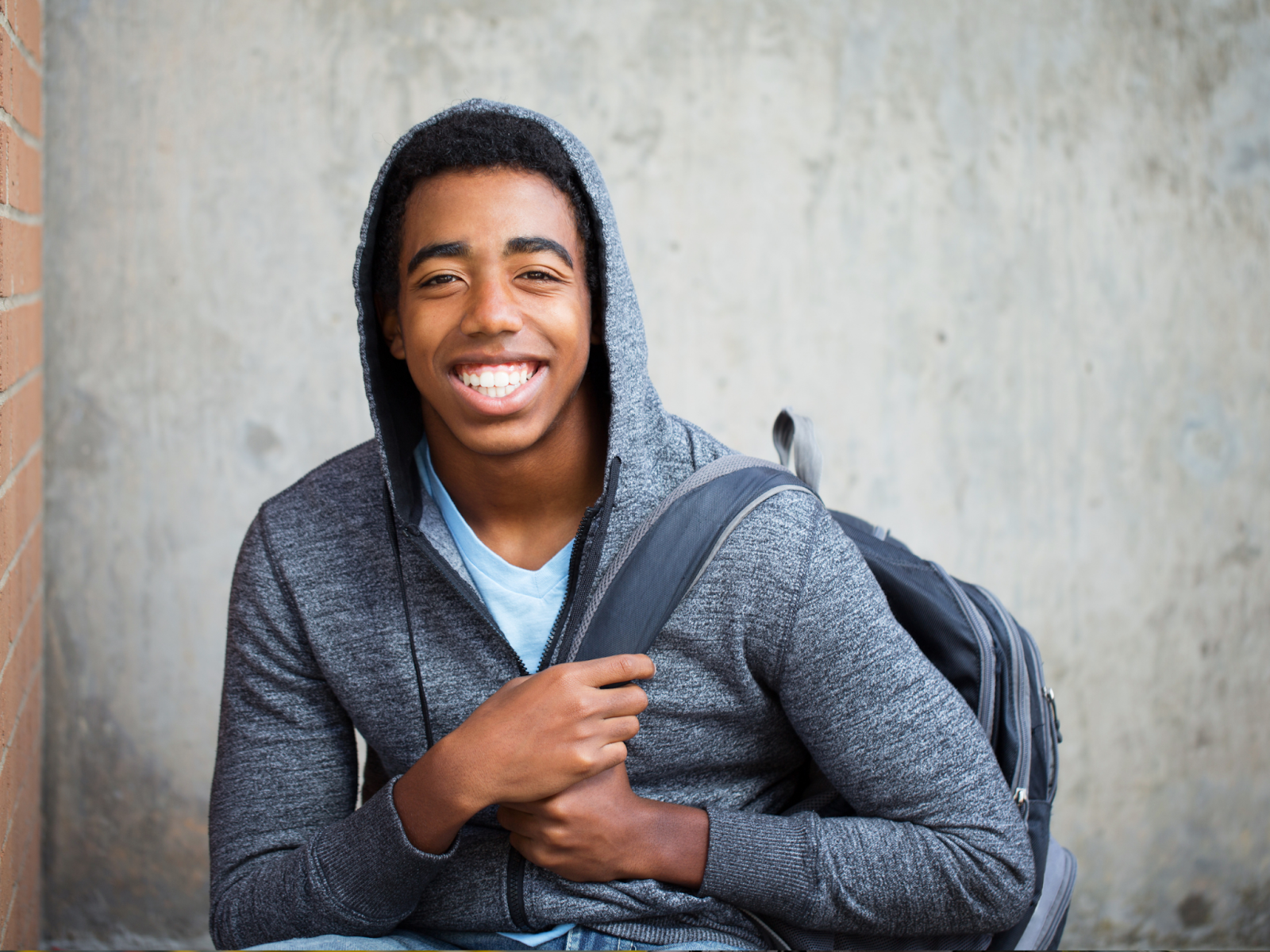 A teenager in a hoodie smiling and holding a backpack.
