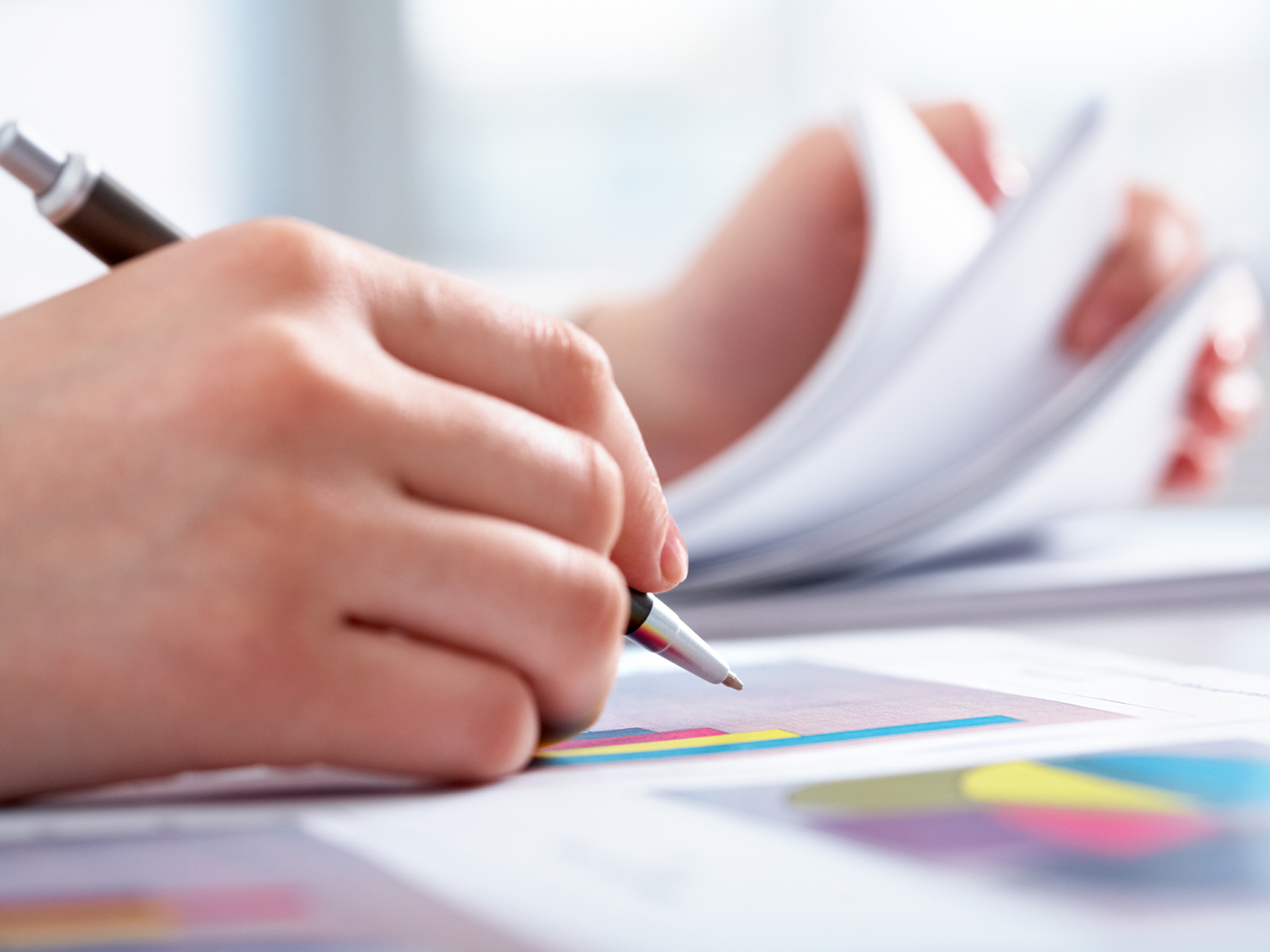 A close-up of a hand holding a pen to paper while flipping through a stack of documents.