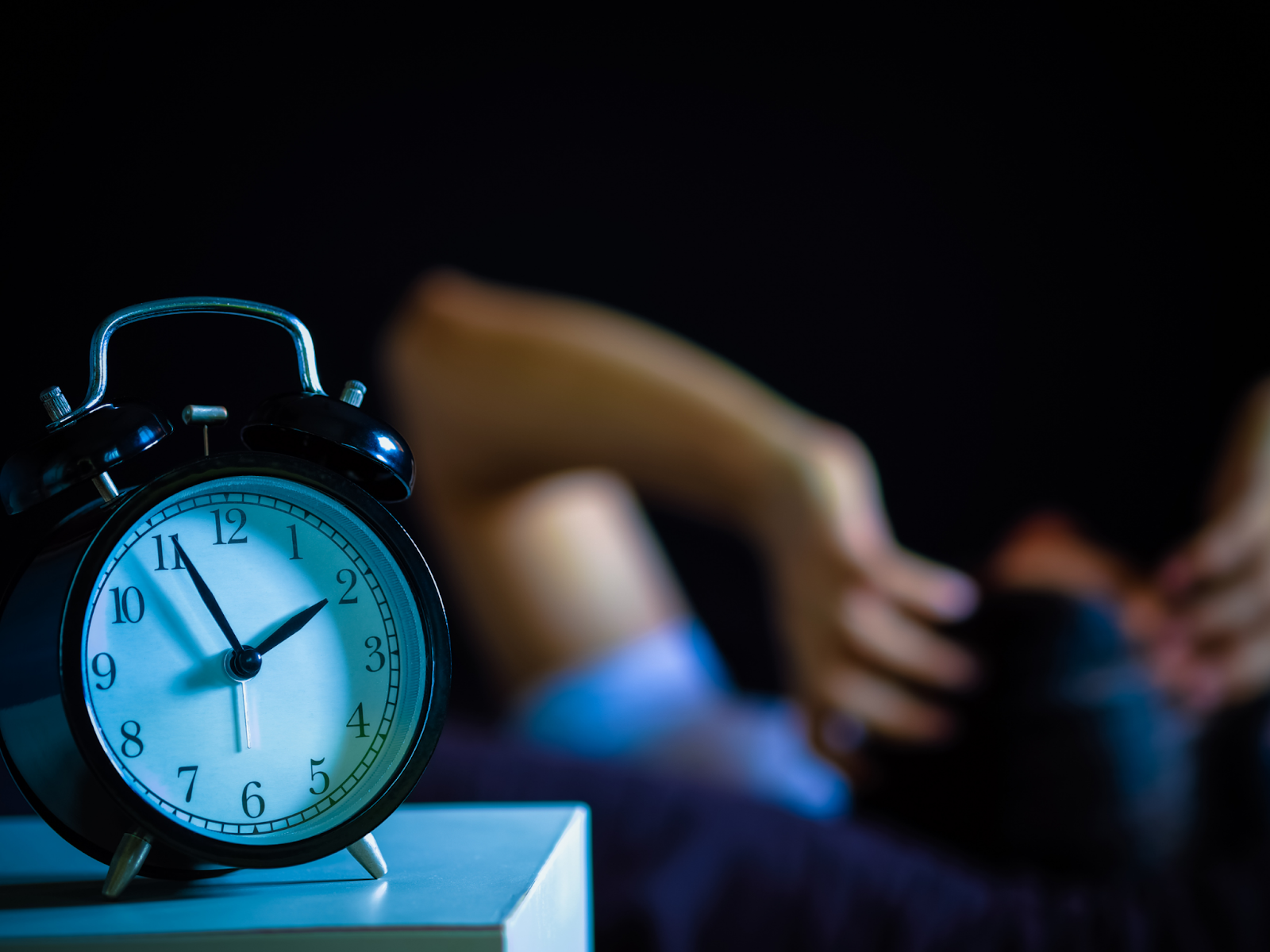 An alarm clock showing the time 1:55 next to a bed where a person has their hands on their head.