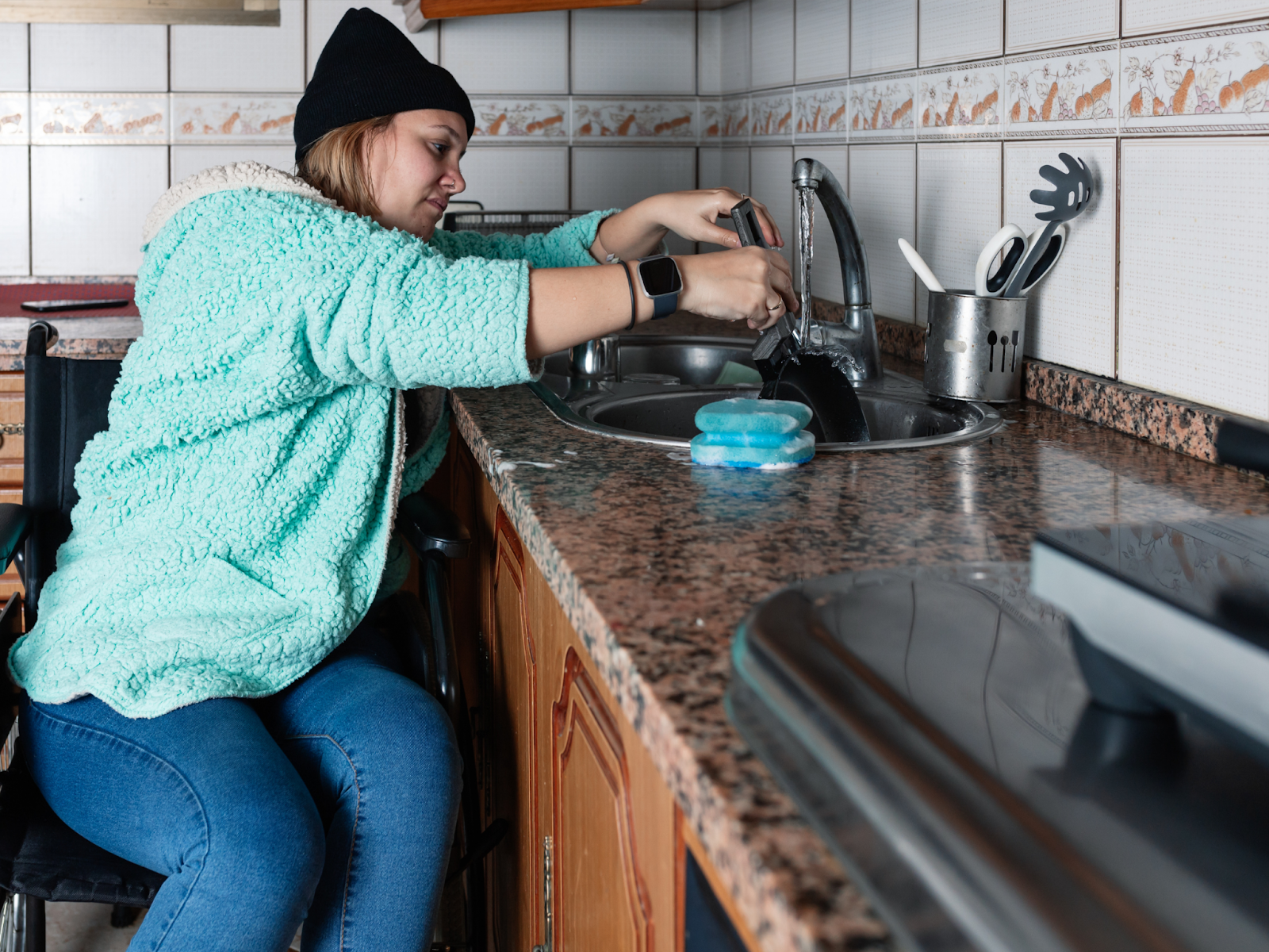 A woman in a wheelchair washing dishes in a kitchen sink.
