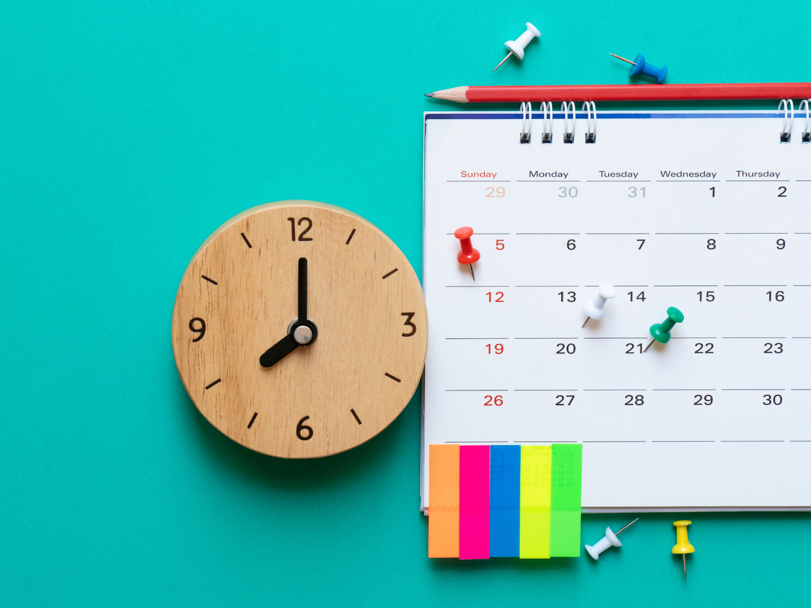 A wooden clock next to a calendar with colored Post-it notes and push pins.