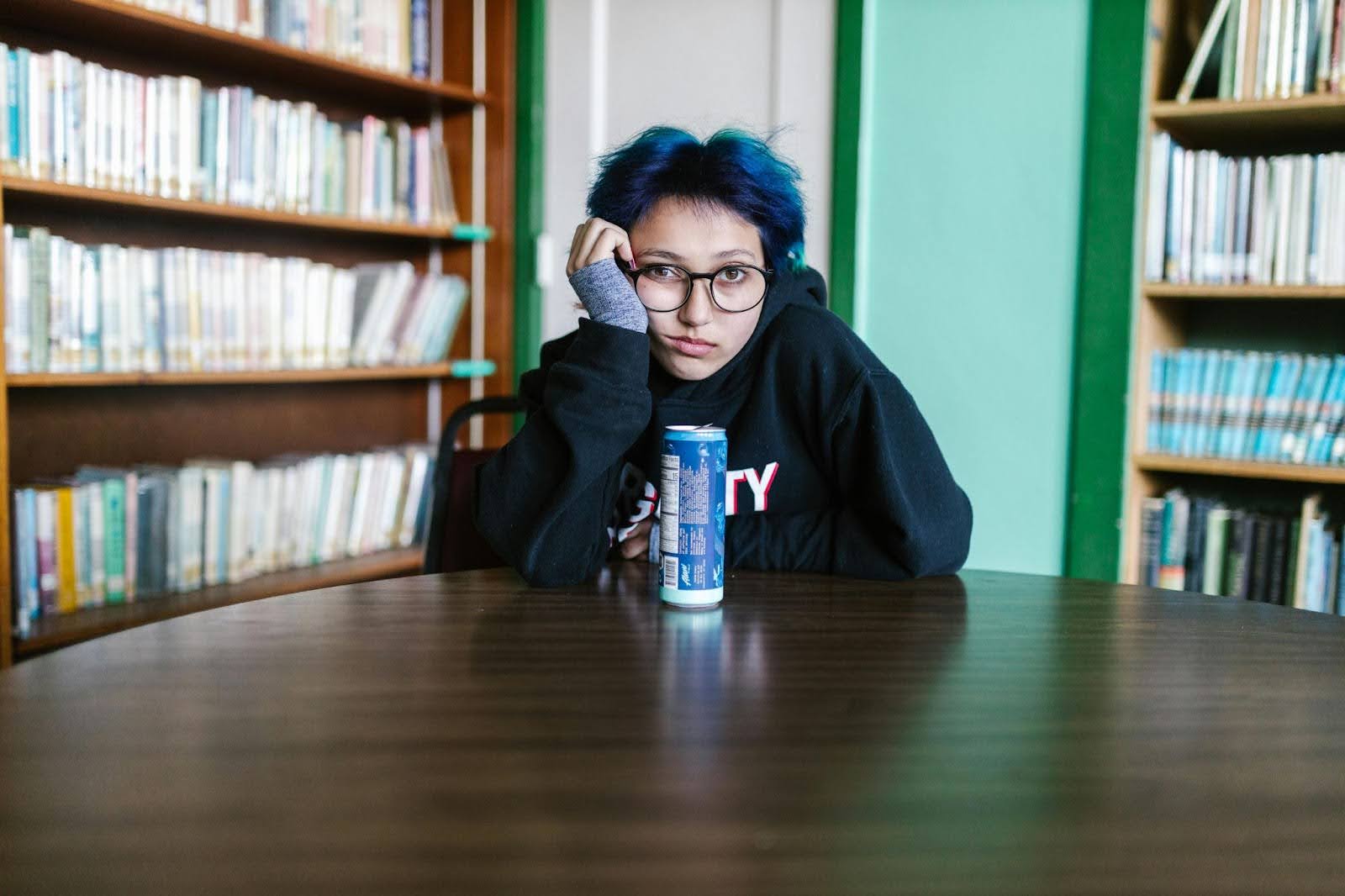A young woman with blue hair sits in a library looking bored and understimulated