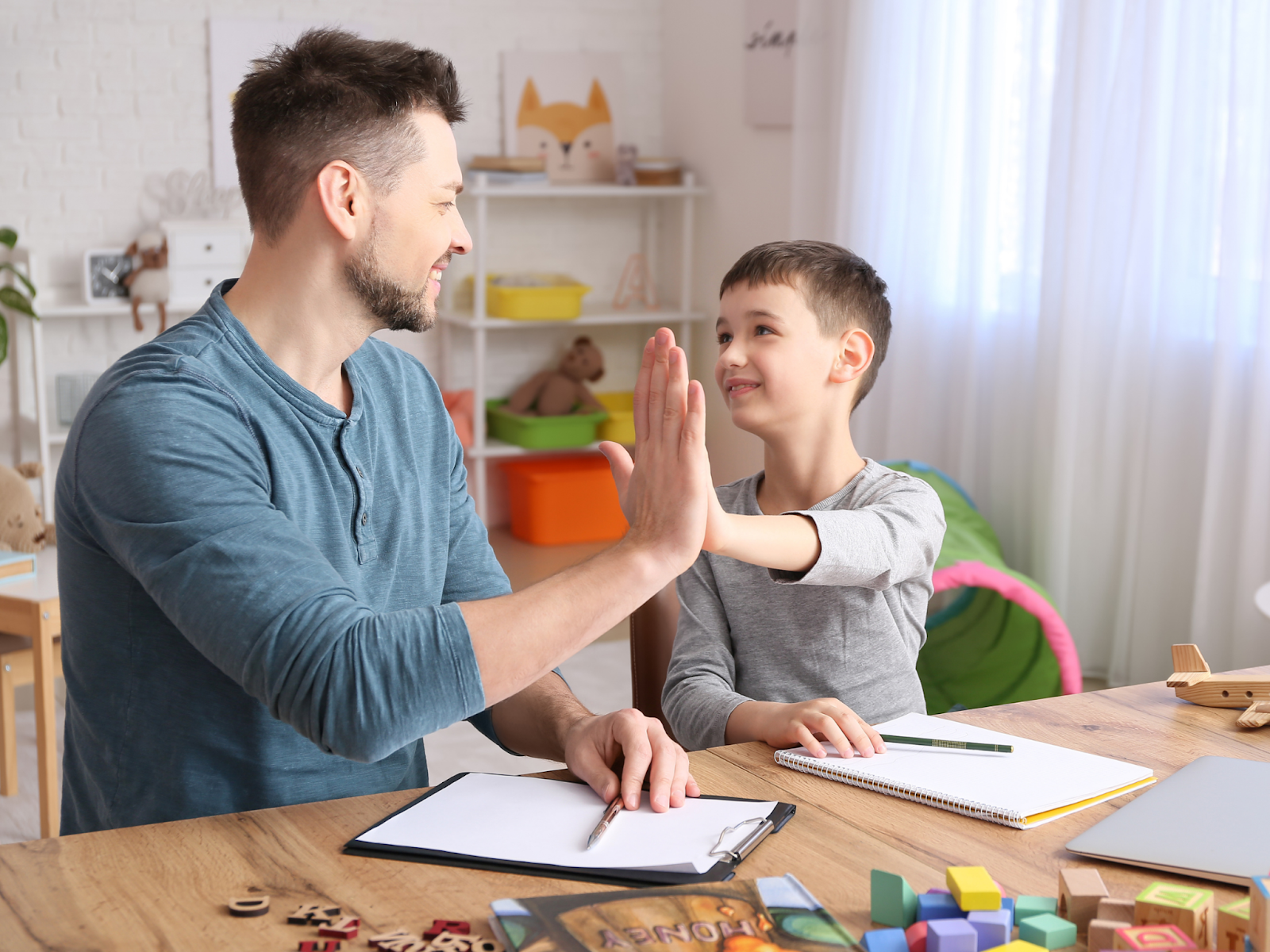 A father and son sit at a table with paper and pens and high-five each other.