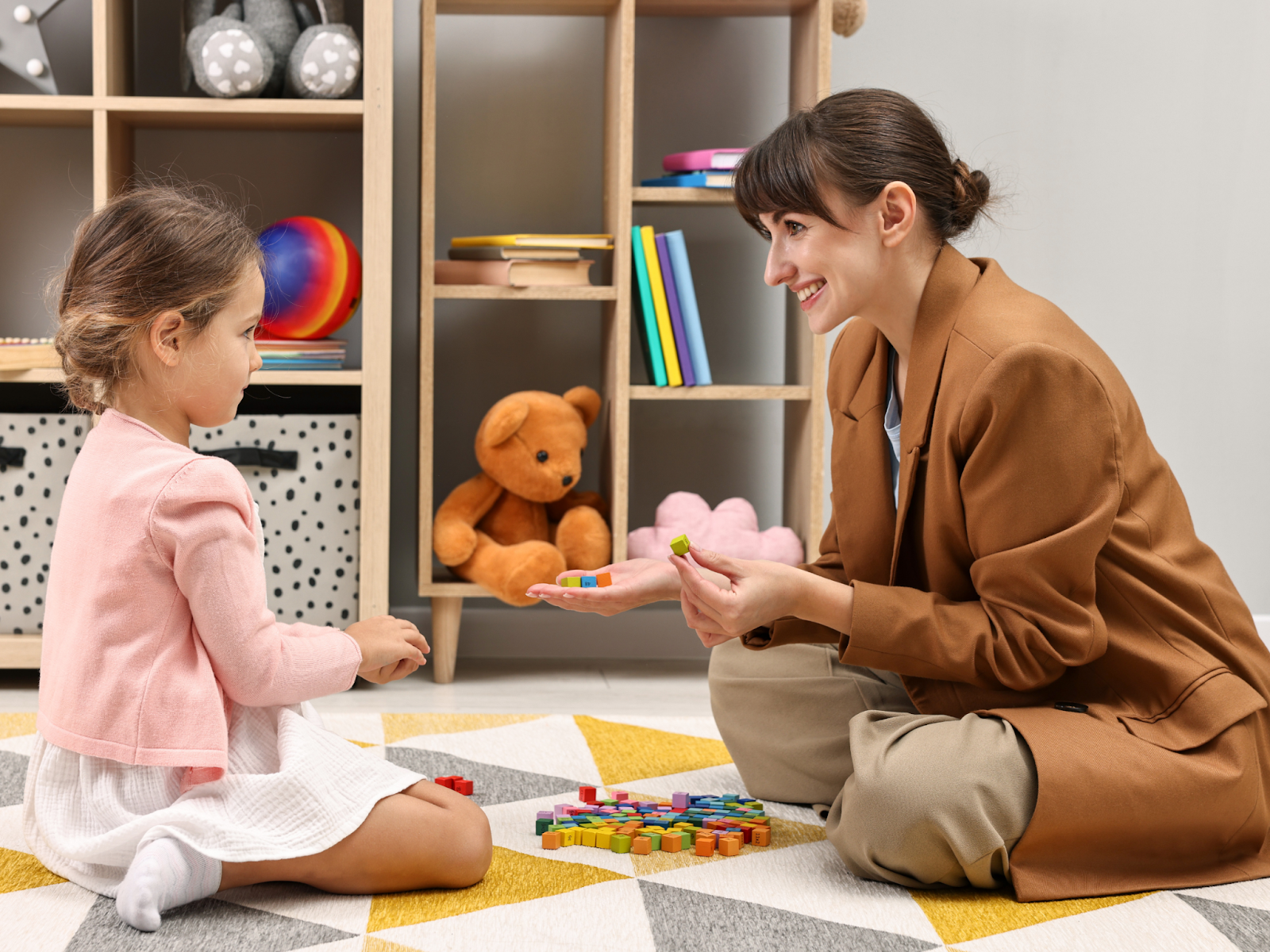 A woman and a young girl sit on a carpeted floor playing with colored blocks.