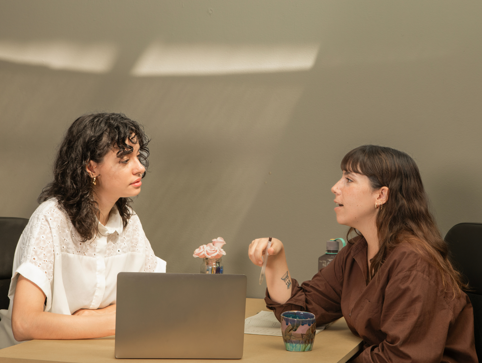 Two women sitting at a table with an open laptop and talking.
