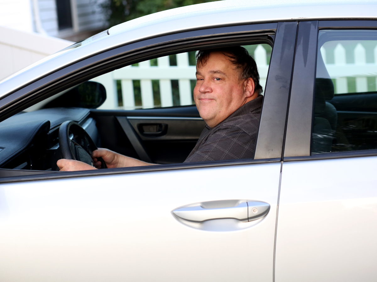 An autistic adult sits in the driver’s seat of a car and smiles while looking out the window