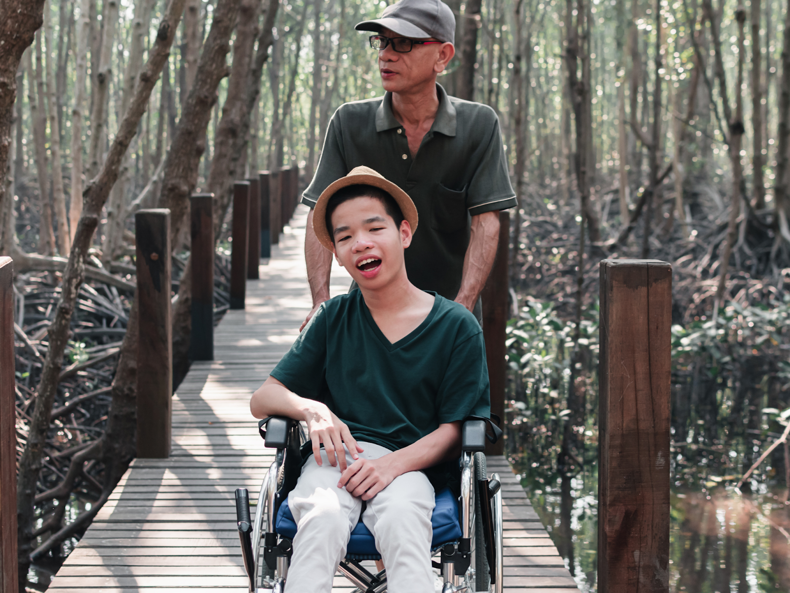 An older man pushing his son in a wheelchair through a wooded area.