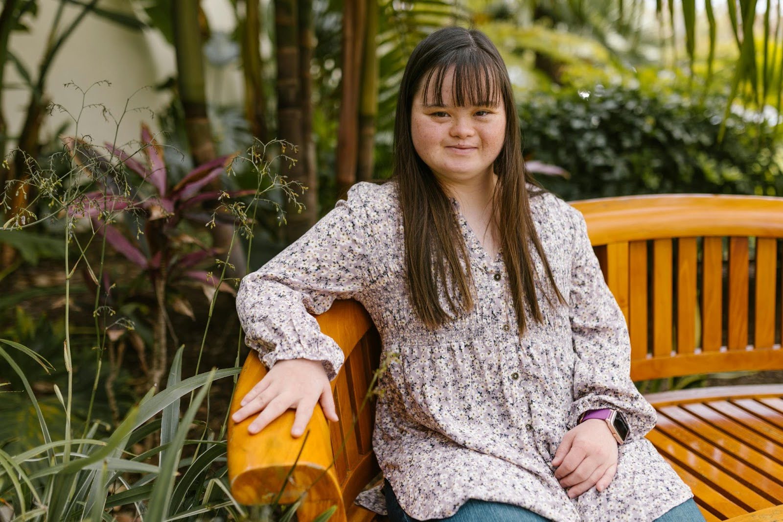 A young autistic woman sits on a wooden bench in a park and smiles.