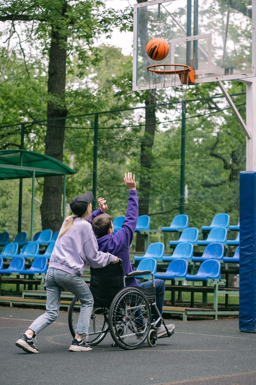 A woman pushes a man in a wheel chair who is shooting basketball