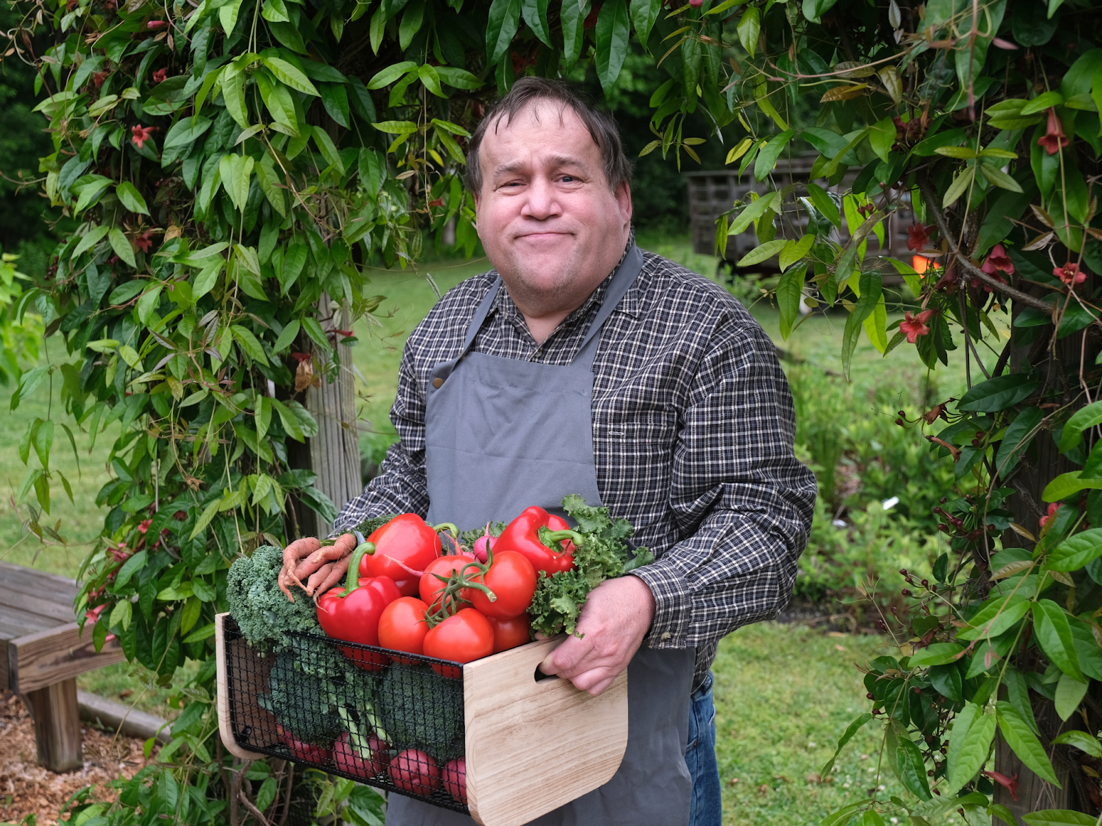 An autistic man standing in his garden holding a crate of tomatoes, bell peppers, and other vegetables.