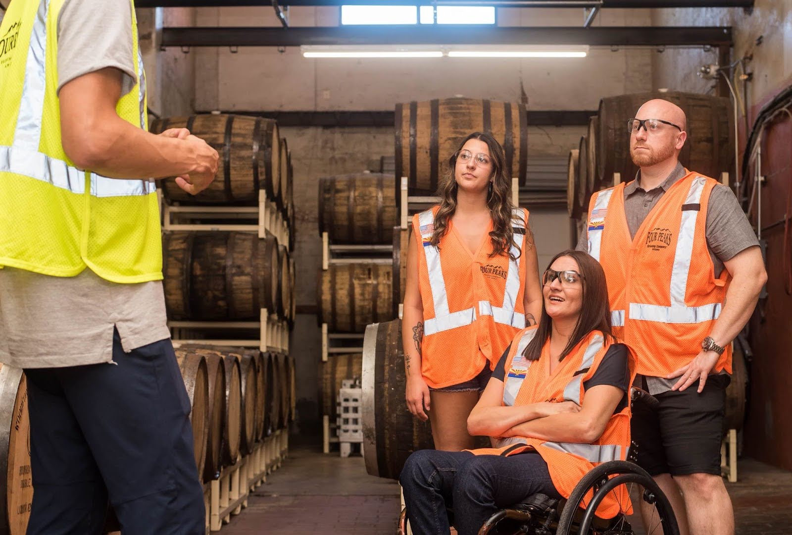 Three people, one in a wheelchair, sit and listen to a supervisor wearing safety goggles and orange reflective vests.