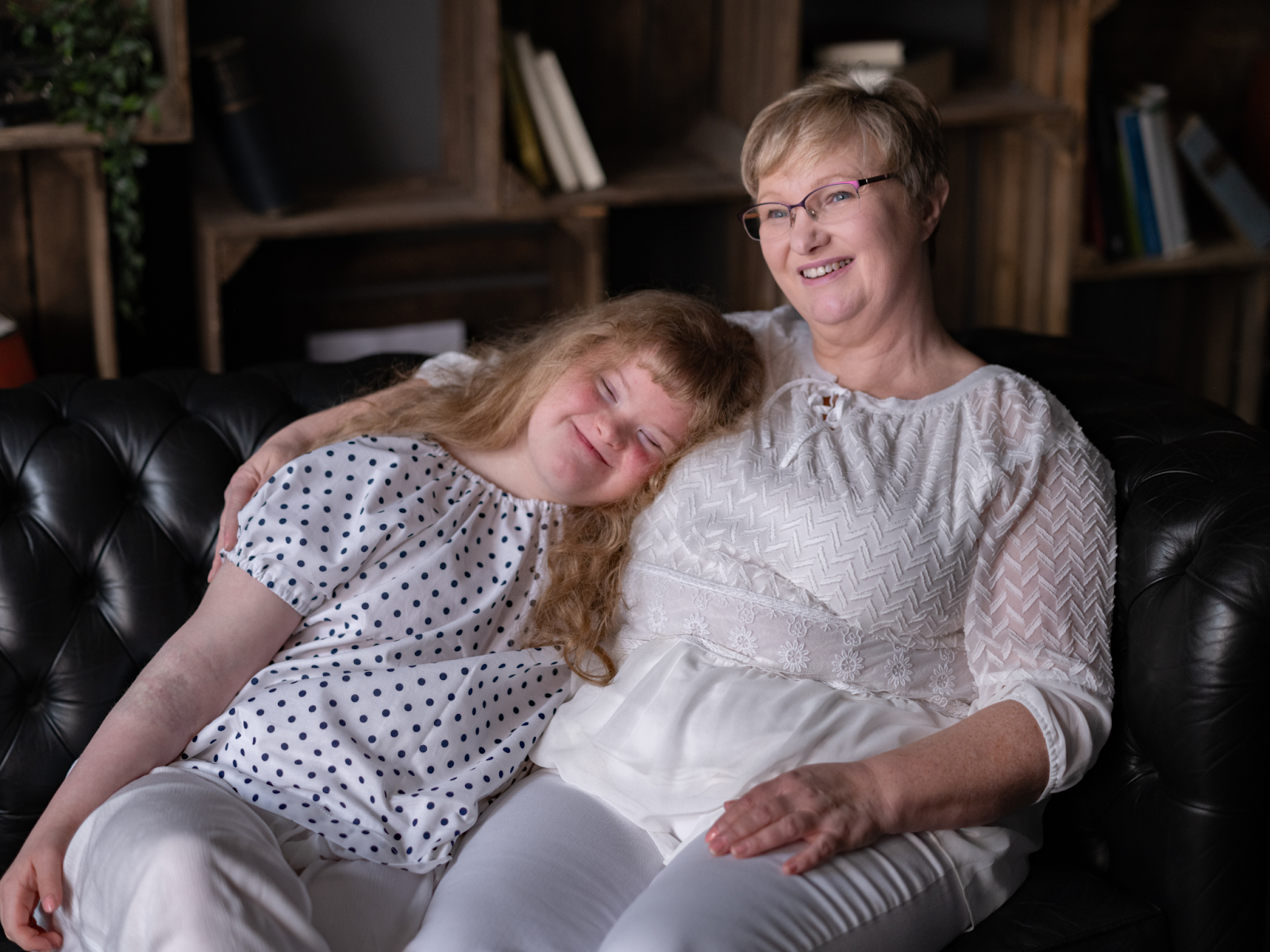 A caregiver and her daughter with Down syndrome sit together on a couch, hugging and smiling.