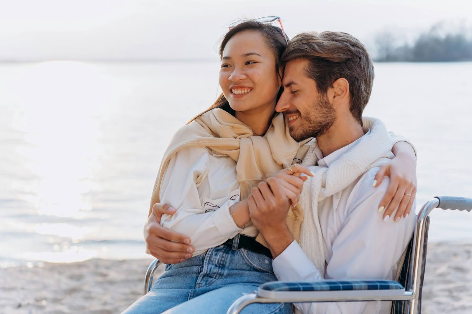 A man in a wheelchair on the beach has a young woman who he loves sitting on his lap