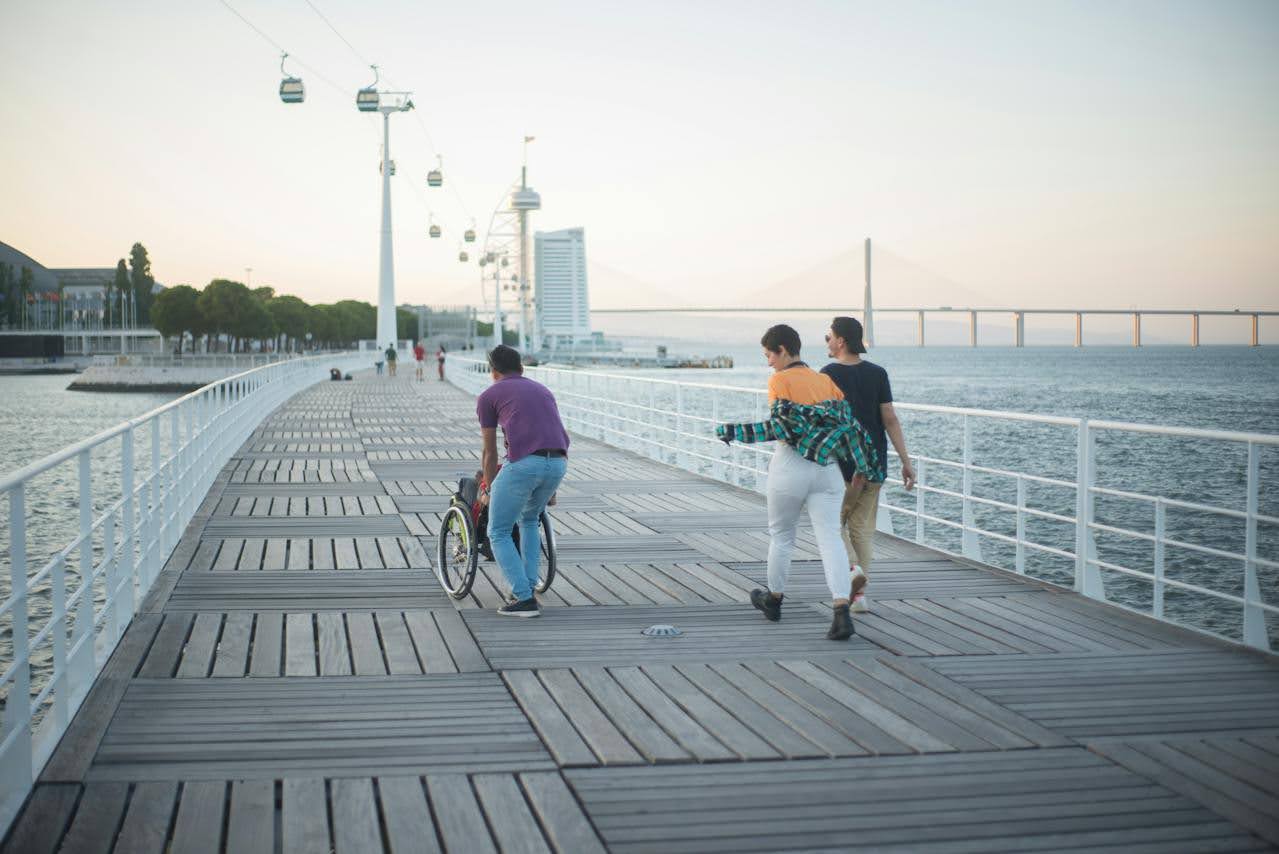 Three friends push a wheelchair on a dock near the ocean.